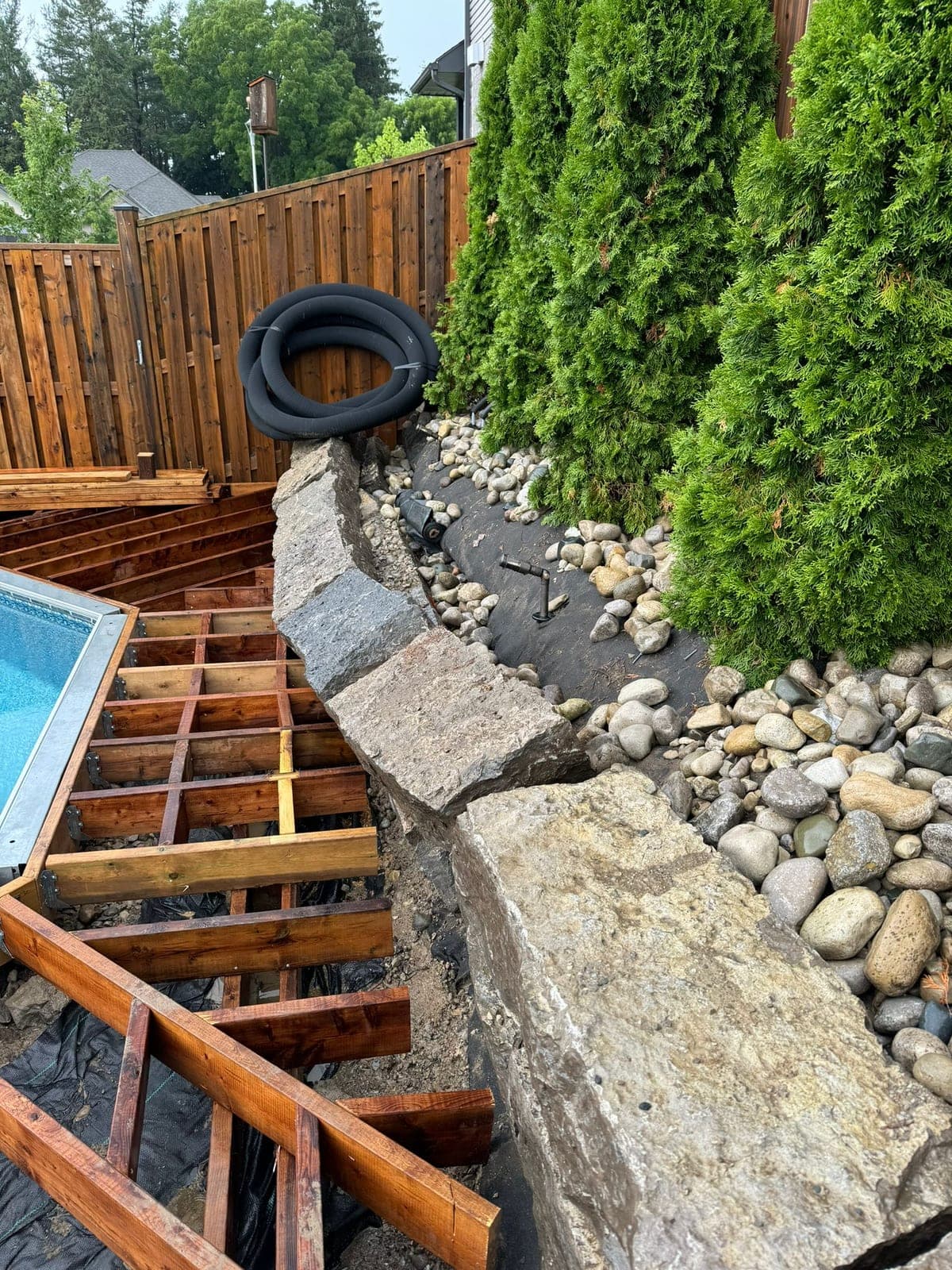 Large boulder retaining wall with river rock drainage bed beside a new backyard pool deck