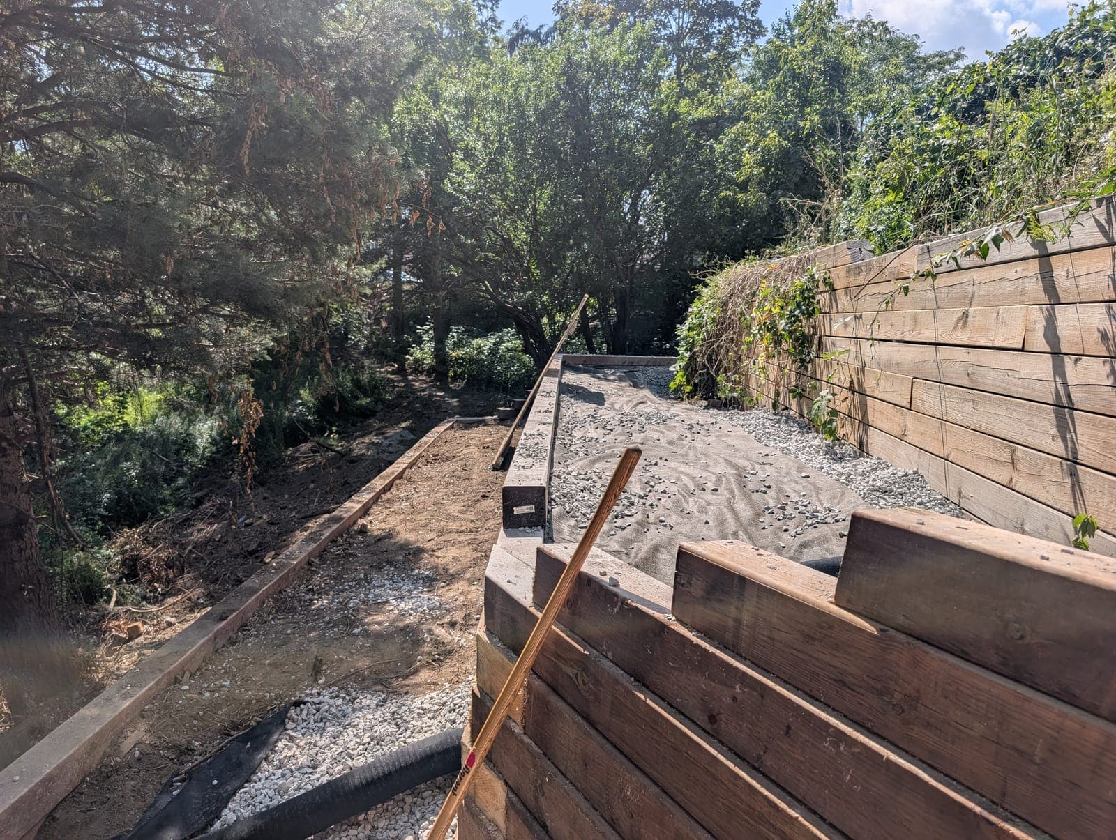 Tall timber retaining wall draped with vines holding back a fenced slope above a gravel patio