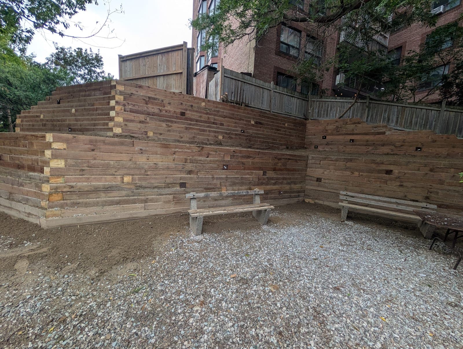 Finished terraced timber retaining wall with built-in bench seating and gravel patio in a shaded backyard
