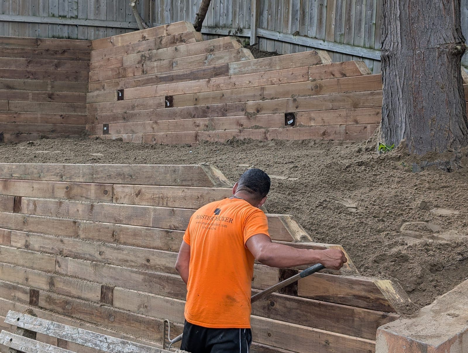 Landscaper grading soil backfill against a tiered timber retaining wall in a backyard