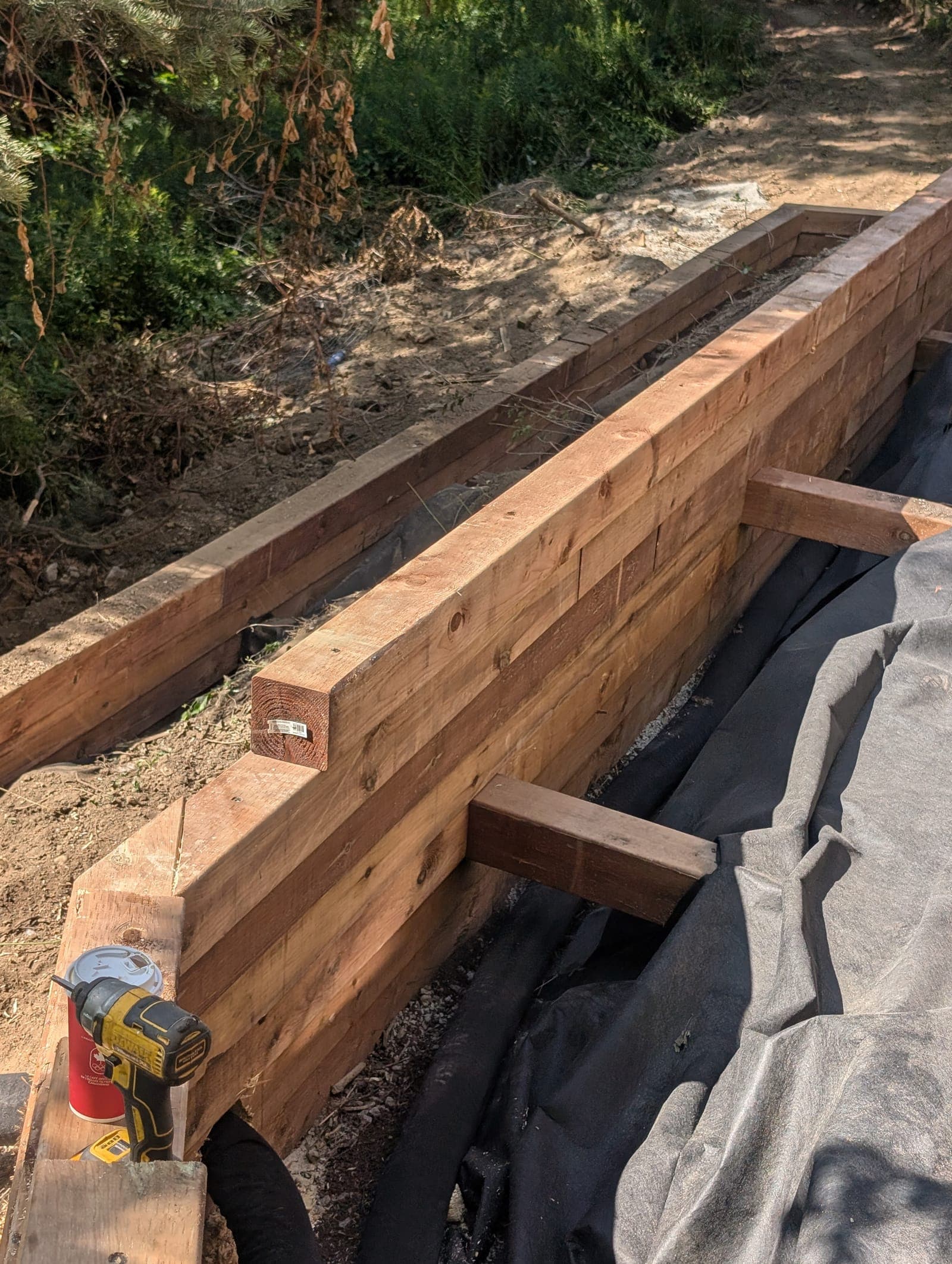 New timber retaining wall under construction with drainage fabric backfill on a wooded backyard slope