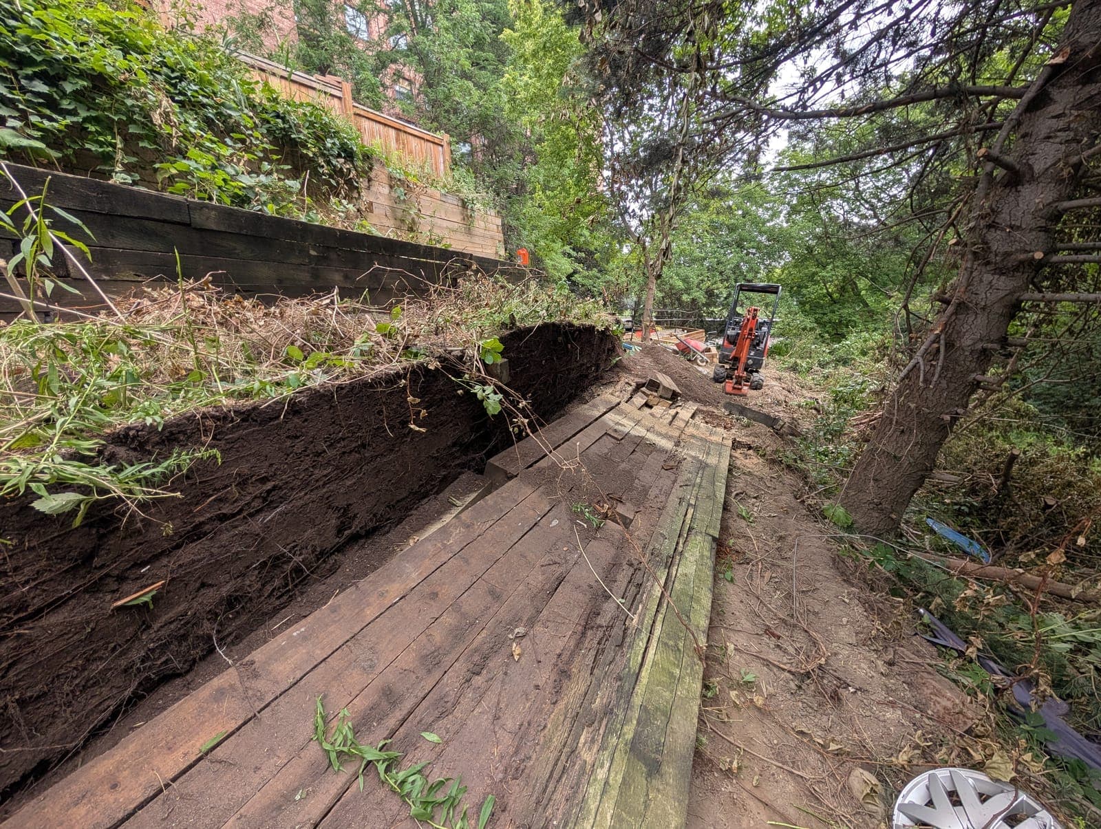 Excavation and site preparation beside an existing timber retaining wall on a wooded slope