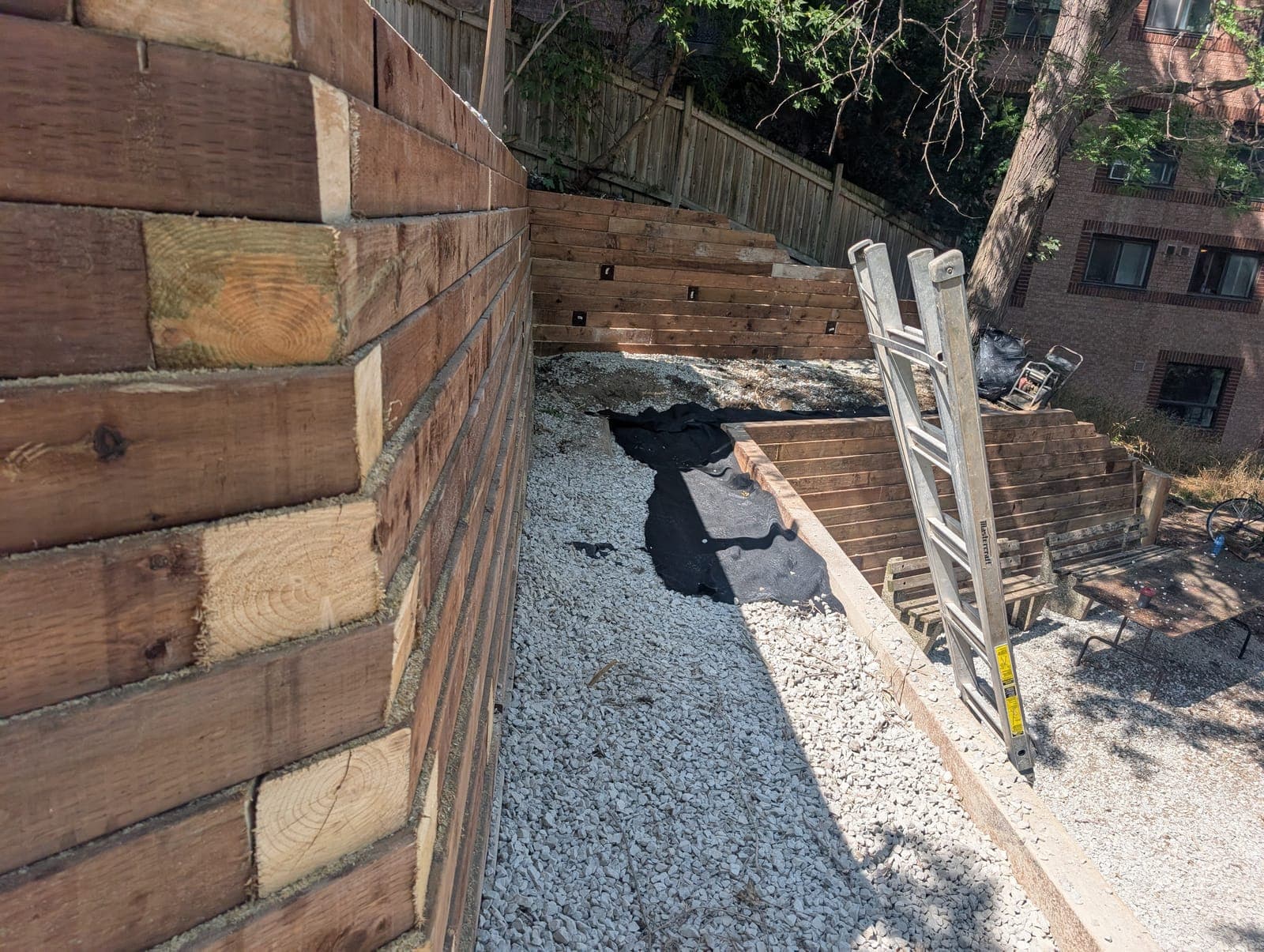 Terraced timber retaining walls under construction with gravel base and drainage fabric in a backyard