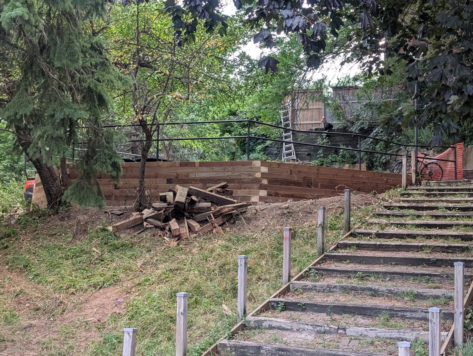 Timber retaining wall with a safety railing beside landscaped stair access on a wooded slope